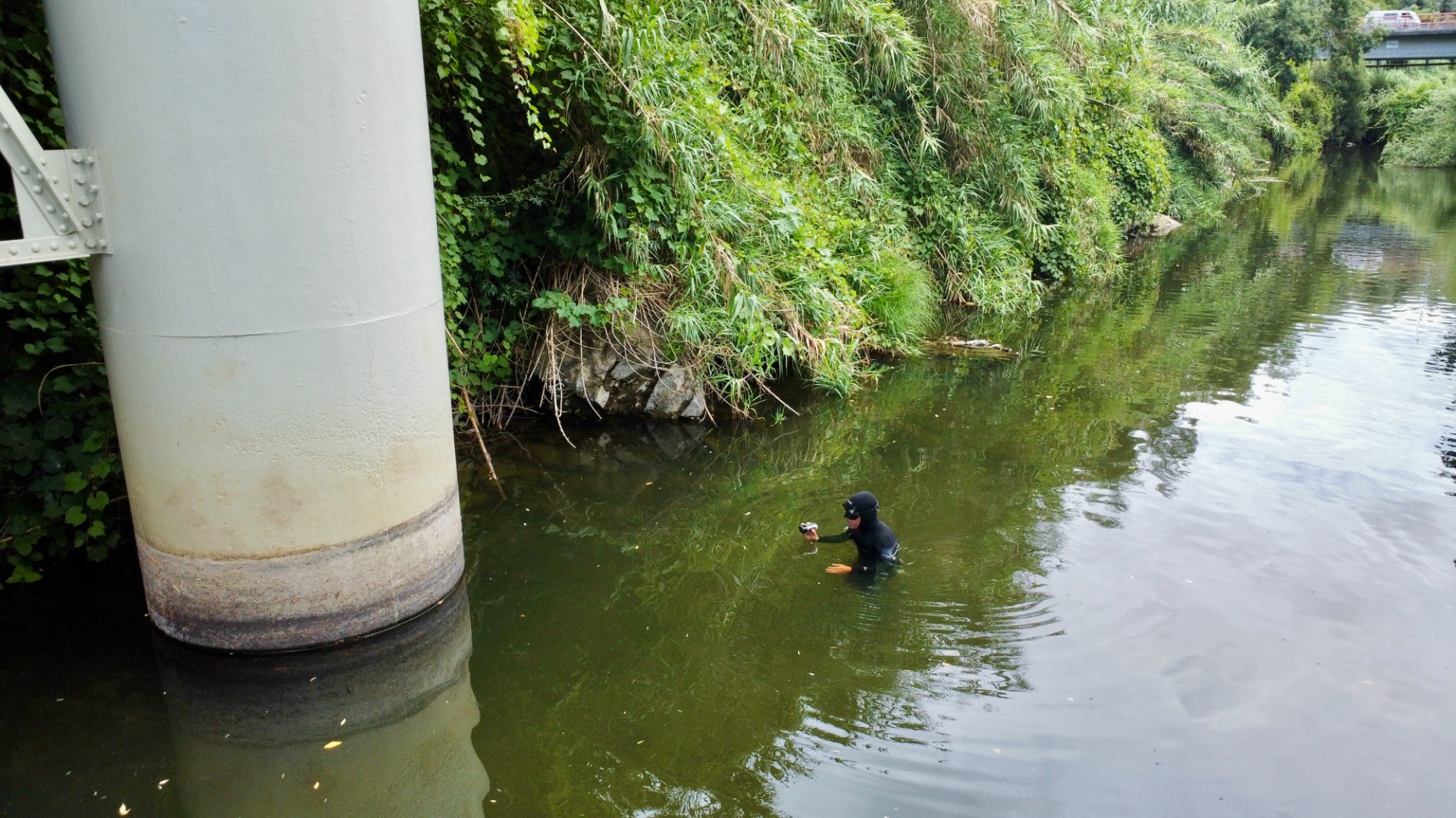 Mergulhador profissional inspecciona em rio verde, pilar de ponte branco e vegetação densa nas margens.