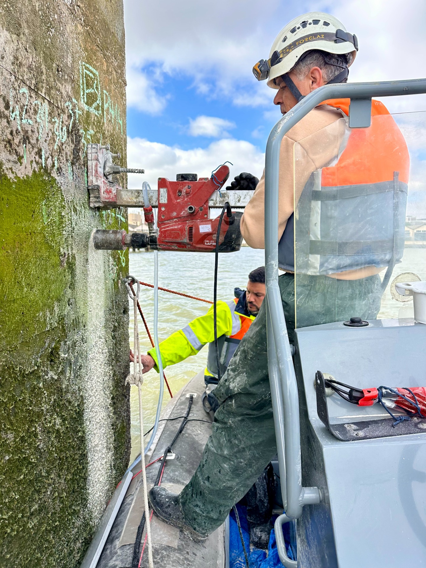 Trabalhadores em barco inspecionam e operam ferramentas em um pilar de concreto, com água ao fundo em Lisboa